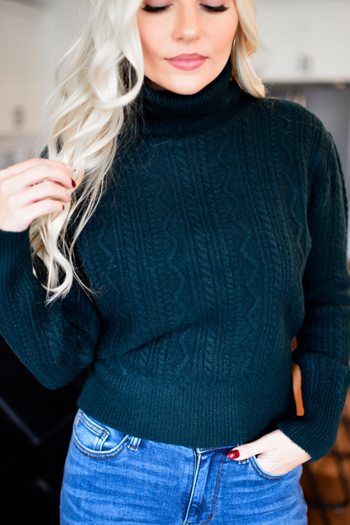 Woman wearing a dark green cable knit sweater and blue jeans indoors.